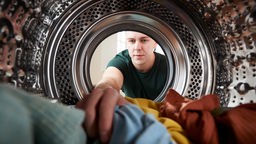 Mann greift in eine Waschmaschine, in der bunte Kleidung liegt. 