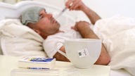 Ein Mann liegt mit Erkältung im Bett, im Vodergrund Tee und Taschentücher