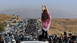 Unverschleierte Frau, die auf dem Dach eines Fahrzeugs steht, während sich Tausende Demonstranten auf den Weg nach Saqez machen.