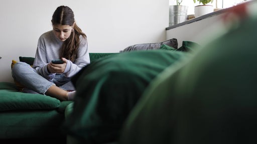Mädchen im Teenager-Alter sitzt auf einer Couch und schaut auf sein Smartphone