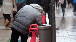Eine Frau durchsucht einen Mülleimer nach Pfandflaschen in einer Fußgängerzone 
