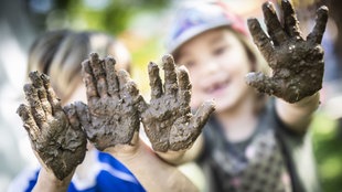 Zwei Kinder zeigen ihre Matschhände im Garten der "Freien Kinderschule" im Stadtteil Unterliederbach. Die Einrichtung existiert seit über 40 Jahren. Angefangen hat alles 1969 in Schwalbach, als Eltern einen der ersten Kinderläden gründeten.