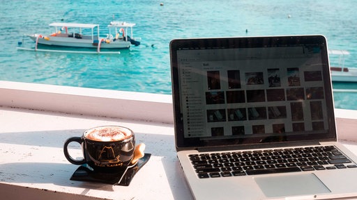 Ein Laptop und ein Kaffee stehen auf einem Tisch, im Hintergrund Meer