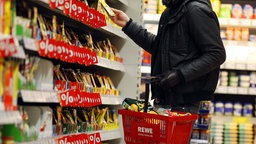 Person steht mit Einkaufskorb in der Hand vor einem Regal im Supermarkt. 