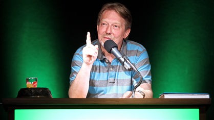 Das Bild zeigt Dietmar Wischmeyer, der an einem Tisch sitzt und den rechten Zeigefinger erhoben hat