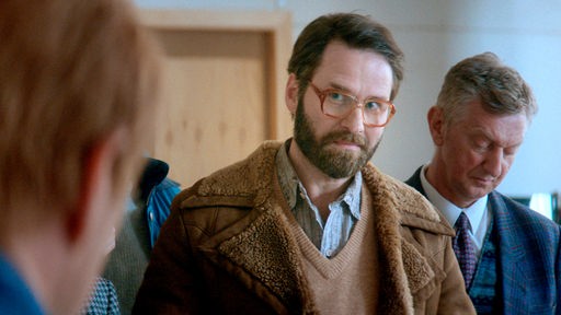 Filmszene aus "Blackport", ein Mann mit Hornbrille und Bart blickt zu einer im gegenüberstehenden Person.