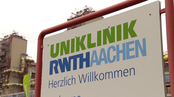 Auf einem Schild steht Uniklinik RWTH Aachen Herzlich Willkommen, dahinter ist ein Gebäude zu sehen.