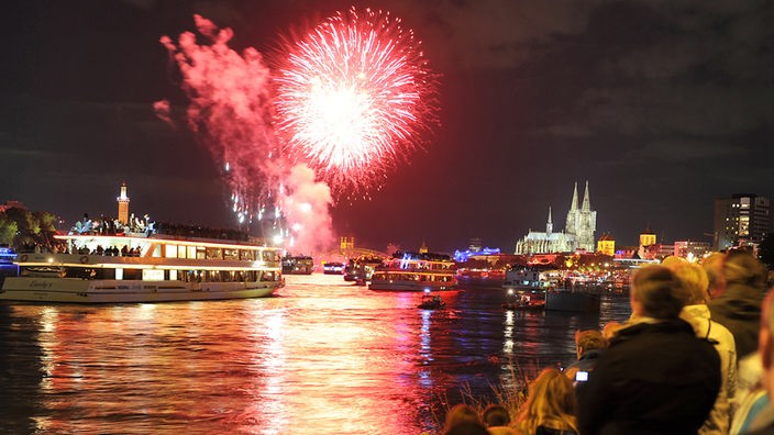 Ein Feuerwerk über dem Rhein in Köln. Auf dem Rhein fahren Schiffe, im Hintergrund ist der Kölner Dom und im Vordergrund sitzende Zuschauer zu sehen