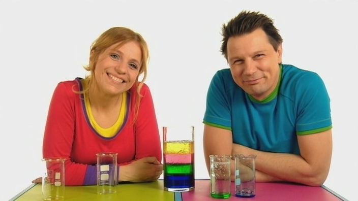 Die Moderatoren stehen neben einem Glas, das mit verschiedenfarbigen Flüssigkeiten gefüllt ist.