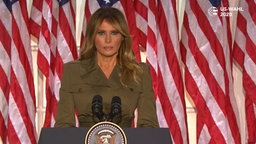 Melania Trump vor US-Flagge bei einer Pressekonferenz, sie trägt einen steifen Armee-Blazer