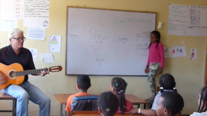 Johannes Remling sitzt mit einer Gitarre vor einer Klasse. Ein Mädchen steht and der Tafel 