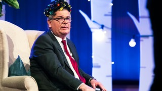 Der Satiriker Jürgen B. Hausmann sitzt auf einem Sofa. Er hat Lockenwickler in den Haaren. 