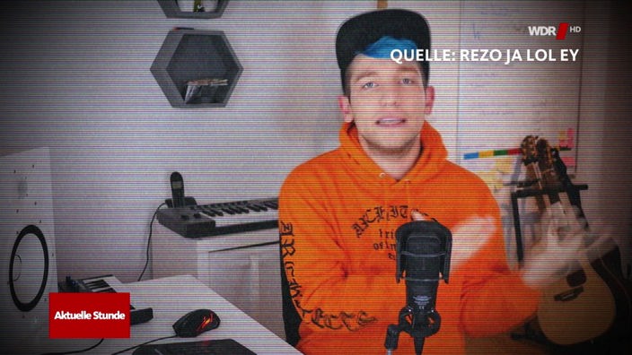 im schwarz gestreiften Bild sitzt ein junger Mann mit orangenem Pullover vor einem Mikrofon
