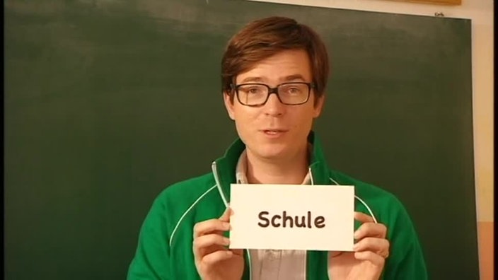 Ralph steht vor einer Tafel in einer türkischen Schule und hält ein Schild vor sich auf dem "Schule" steht.