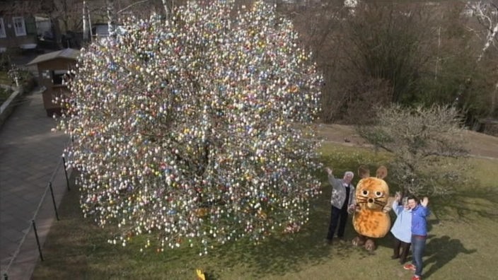 Andre, die Maus und ein Ehepaar stehen neben einem Baum, der mit Ostereiern gescgmückt ist