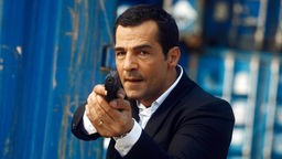 Kommissar Mehmet Özakin (Erol Sander) zielt mit einer Pistole, hinter ihm eine Wand von blauen Hafencontainern.