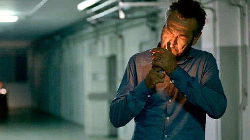 Ein Mann steht in einem grauen Kellerflur und zündet sich eine Zigarette an.