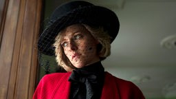 Eine Frau mit schwarzem Hut und Netz vor dem Gesicht blickt betrübt aus dem Fenster