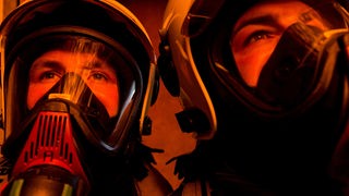 Zwei Feuerwehrmänner mit Atemschutzmaske und Helm in rotem Licht.