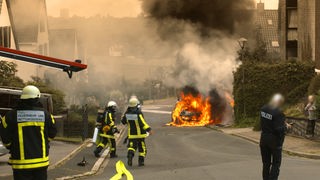 In einem Wohngebiet brennt es auf der Straße, Einsatzkräfte der Feuerwehr und der Polizei sowie ein Anwohner befinden sich in der Nähe des Feuers. 