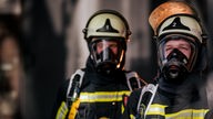 Zwei Feuerwehrmänner mit Helm und Atemmaske. Einer der beiden steht im Hintergrund.