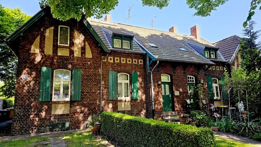Ein Fachwerkhaus mit grünen Fensterläden und kleinem Vorgaretn, das im Schatten grüner Bäume steht. Die Arbeitersiedlung Rheinpreußen im heutigen Duisburg Homberg.