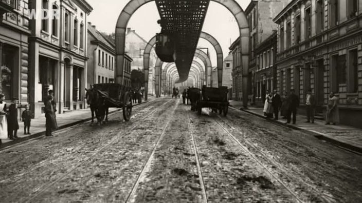 Alte Foto der Wuppertaler Schwebebahn über einer Straße mit Pferfuhrwerken