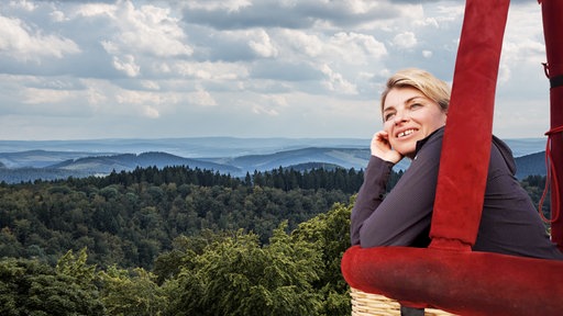 eine blonde Frau in einem Heißluftballon blickt nachdenklich zum Himmel, im Hintergrund unter ihr ein Wald