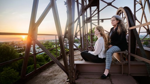 Zwei Frauen sitzen im Licht der untergehenden Sonne vor industrieller Kulisse 
