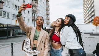 Drei hübsch gestylte Mädchen schießen ein Selfie mit dem Handy und lachen/ lächeln.