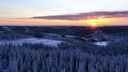 vorne verschneiter Nadelwald bei Dämmerlicht, dahinter Hügel und Wälder teils mit Schnee, knapp über dem Horizont die orange Sonne und oranger Himmel mit Schleierwolken