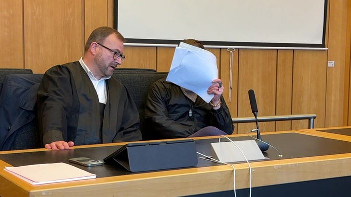 Die angeklagte Person verdeckt ihr Gesicht mit Blättern Papier. Neben der Person ist sitzt ein Anwalt.