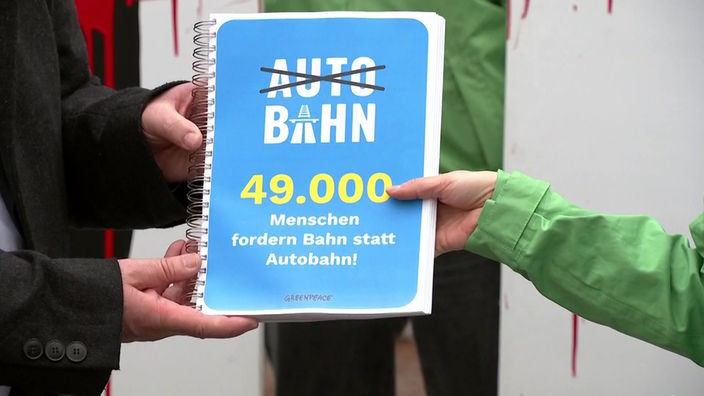 Zwei Personen halten eine Petition in den Händen: Dort steht "AUTO" durchgestrichen und dadrunter nicht durchgestrichen "BAHN 49.000 Menschen fordern Bahn statt Autobahn! GREENPEACE".