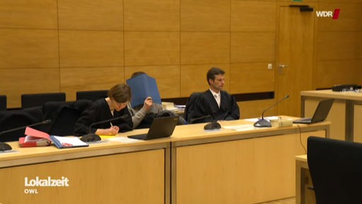 In einem Gerichtssaal sitzen zwei Anwälte in schwarzer Robe und in der Mitte sitzt ein Mann, der sich eine blaue Mappe vor das Gesicht hält.