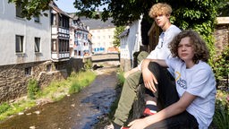 Zwei Kinder sitzen am Rand eines Flusses, der mitten durch eine Stadt in der Eifel führt.