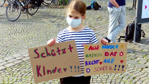 Ein Kind mit Mundschutz bei einer Kundgebung: Das Mädchen hält zwei Plakate hoch mit den Aufschriften "Schützt Kinder" und "Alle Kinder haben das Recht auf Schutz"