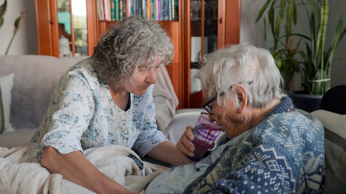 Eine grauhaarige Frau sitzt in einem Wohnzimmer links neben einer weißhaarigen, älteren Frau und hilft ihr beim Trinken.