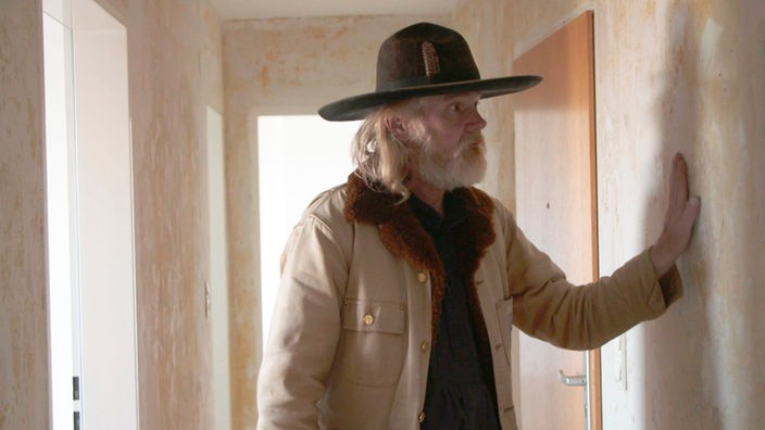 Ein Mann mit Hut berührt die Wand in einer leeren Wohnung