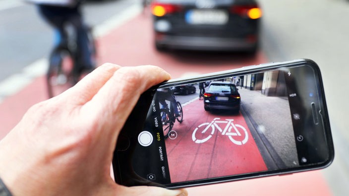 Blick auf ein  Smartphone-Display:  Ein Kfz parkt auf dem Radweg; ein:e Radfahrer:in ist gezwungen, auszuweichen 