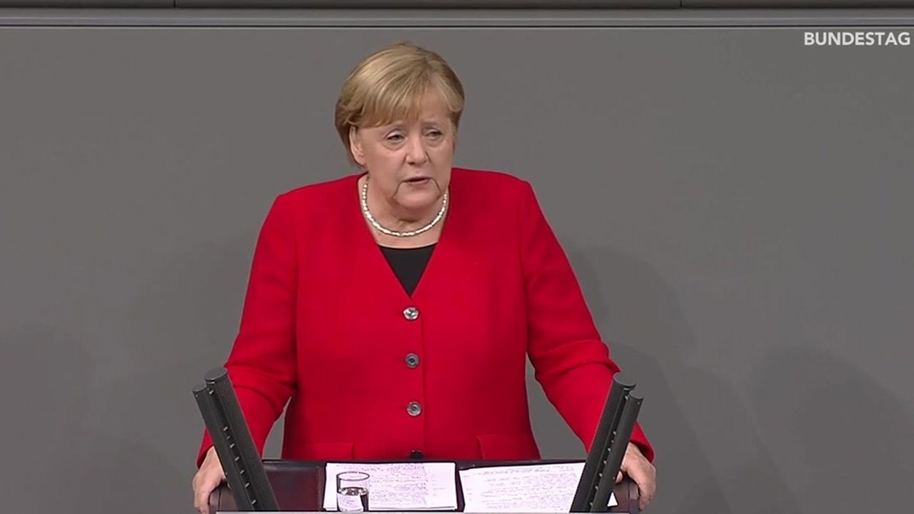 Angela Merkel steht an einem Rednerpult. Vor ihr steht ein Glas Wasser und Notizblätter. Sie stützt sich mit beiden Armen auf das Pult.