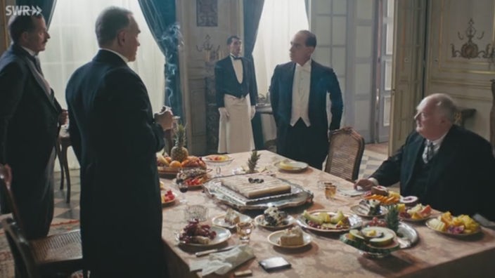Filmszene: Drei Männer stehen um einen gedeckten Tisch, ein Mann sitzt
