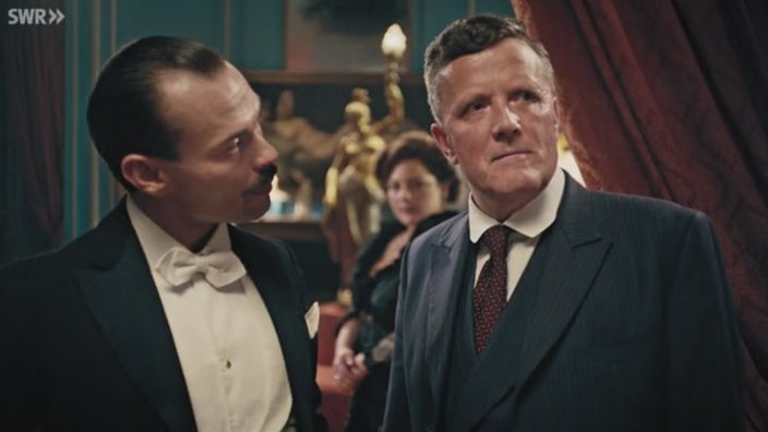 Filmszene: Zwei elegant gekleidete Männer im Gespräch