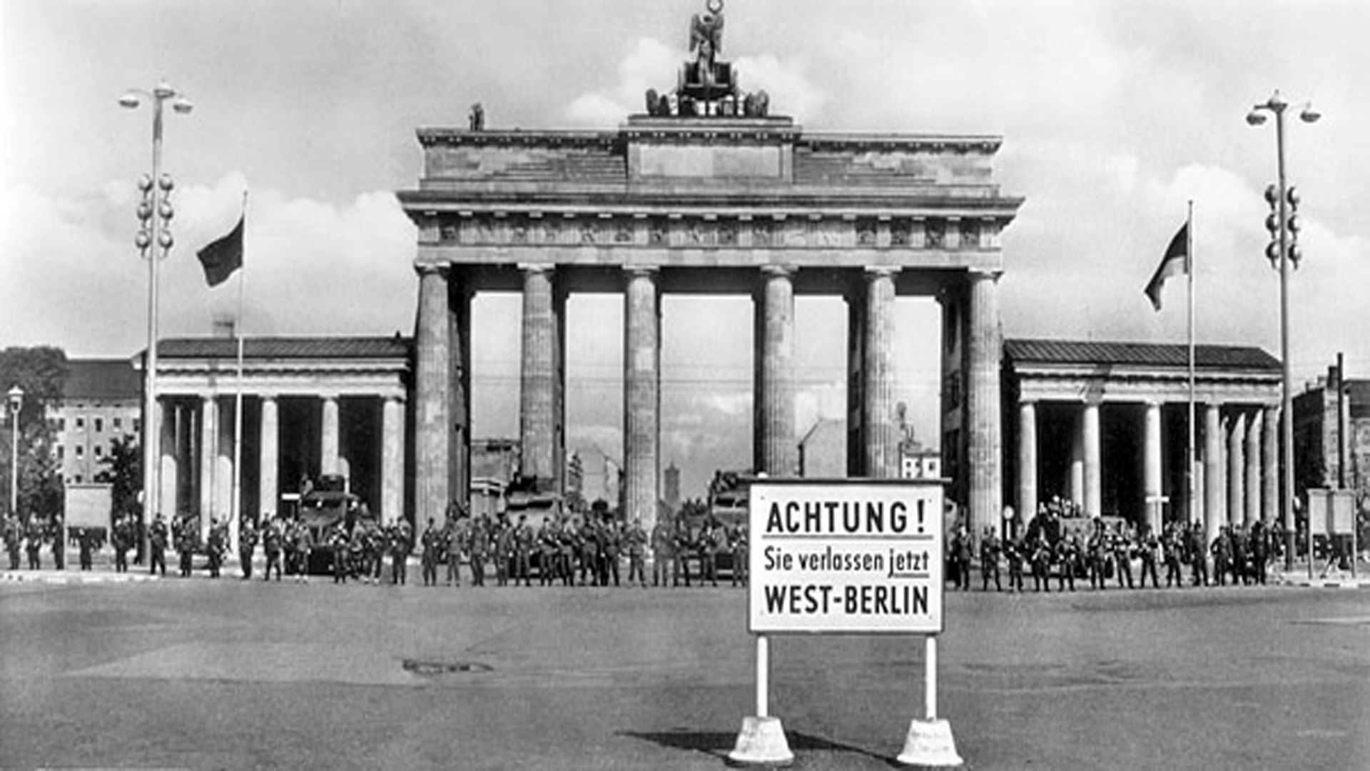 Schwarzweiß-Bild: Brandenburger Tor mit Schild "Achtung! Sie verlassen jetzt West-Berlin" davor.
