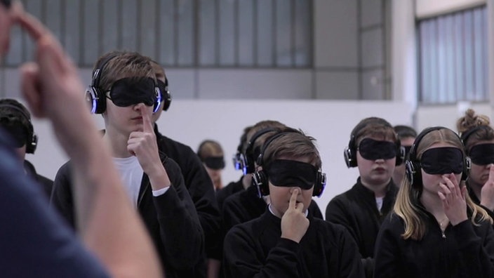 In einer Halle steht eine Gruppe von Kindern mit verbundenen Augen und Kopfhörer auf, die sich gerade an die Nase fassen.