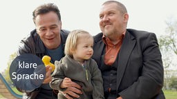 Die Kommissare Freddy Schenk (Dietmar Bär) und Max Ballauf (Klaus J. Behrendt) kümmern sich liebevoll um ein Kind. Max Ballauf hält dem kleinen Mädchen eine gelbe Gummiente hin, während es lächelnd auf Freddy Schenks Schoß sitzt.
