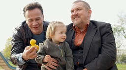 Die Kommissare Freddy Schenk (Dietmar Bär) und Max Ballauf (Klaus J. Behrendt) kümmern sich liebevoll um ein Kind. Max Ballauf hält dem kleinen Mädchen eine gelbe Gummiente hin, während es lächelnd auf Freddy Schenks Schoß sitzt.