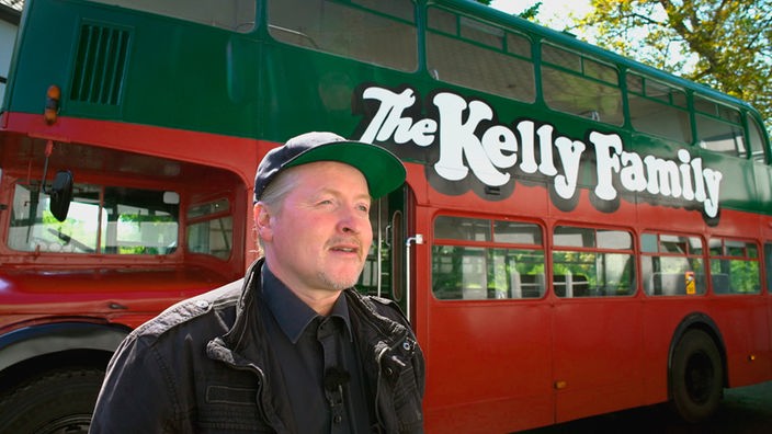 Musiker Joey Kelly steht vor dem roten Bus mit dem großen Schriftzug "Kelly Family".