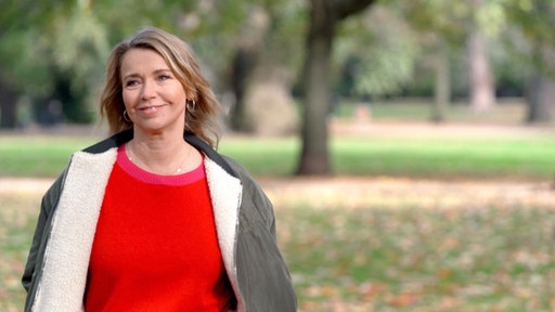 Tina Ruland in rotem Pullover und mit Jacke darüber spaziert im Park.