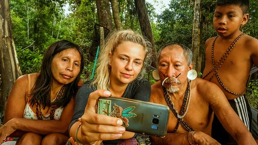 Eine Frau filmt sich und einen indigenen Mann und eine indigene Frau und ihren Sohn auf einer Lichtung im Amazonas