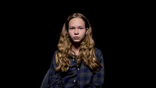 Eine junge Frau steht vor einem dunklen Hintergrund und blickt entschlossen in die Kamera